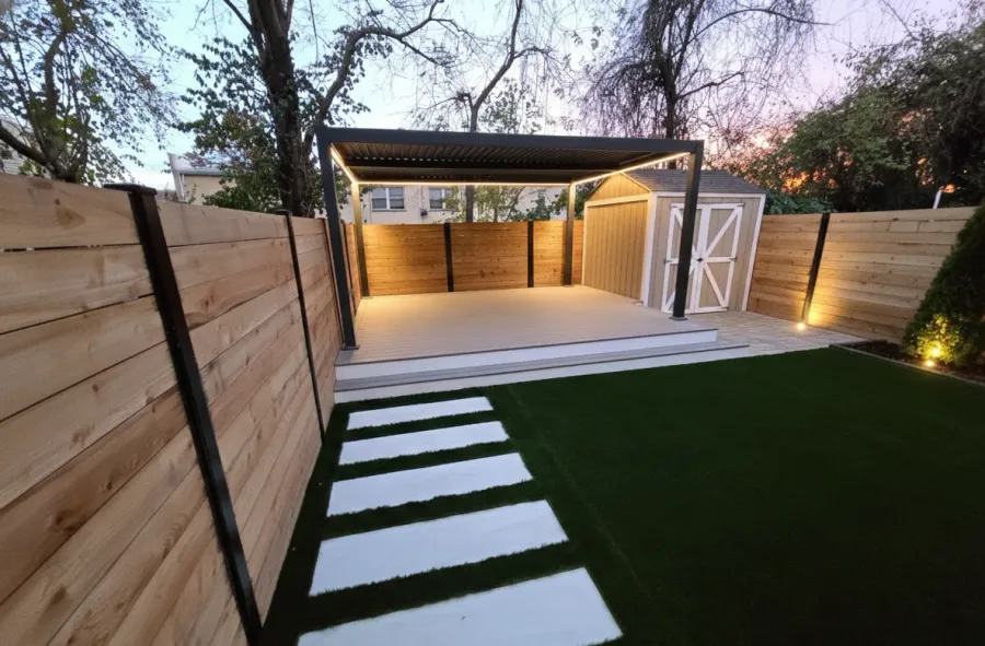 Completed backyard at dusk with lit pergola, shed, stepping-stone path and artificial turf